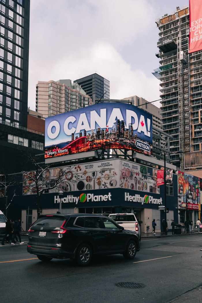 A bustling street in Toronto with a prominent O Canada billboard, cars, and skyscrapers.